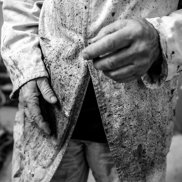 Les mains de Noël Dolla sur sa blouse d’atelier tachée de peinture, en noir et blanc